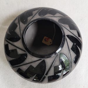 May include: A round, black and gray ceramic bowl with a central opening. The bowl features a geometric design of black shapes on a gray background. A small tag is attached to the inside of the bowl.