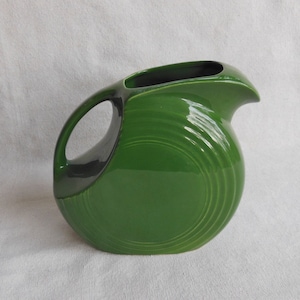 May include: A green ceramic pitcher with a wide, rounded base and a curved spout. The pitcher has a smooth, glossy finish and a unique, swirling design.