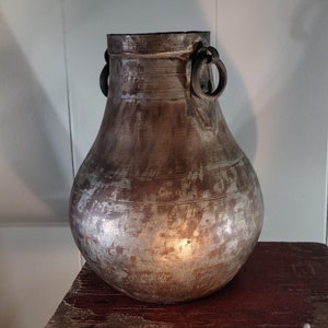 May include: A large, round, silver-colored metal vase with two iron handles. The vase has a worn, aged appearance and is sitting on a wooden surface.