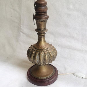 May include: A brass table lamp with an ornate design and a wooden finial. The lamp has a round base and a cord with a switch.