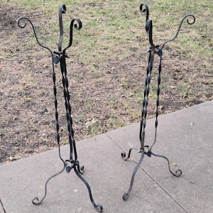 May include: Two black wrought iron plant stands. Each stand has a twisted central column, three curved legs, and decorative scrollwork at the top and base. The stands are displayed outdoors on a concrete surface.