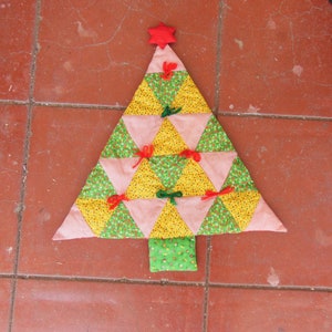 May include: A handmade Christmas tree decoration. The tree is constructed from colorful fabric triangles in green, yellow, and red, with small bows and a red star at the top. The tree is placed on a red tile surface.