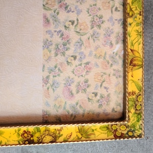 May include: A vintage picture frame with a yellow floral border and a floral patterned background. The frame has a gold rope trim and a cream-colored background on the left side. The floral pattern includes pink, yellow, and blue flowers.