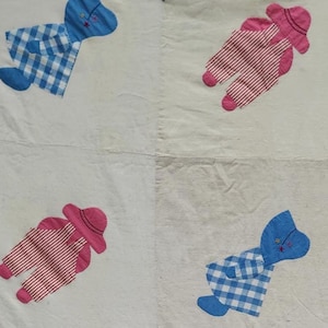 May include: A vintage quilt with a white background featuring appliqued figures of babies in pink and blue. The babies are wearing hats and overalls. The quilt has a pink border and blue heart accents.