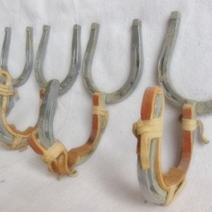 May include: A collection of silver-colored horseshoe-shaped wall hooks. Each hook has a leather and woven accent in shades of tan and brown. The hooks are arranged against a white background.