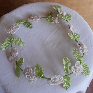 May include: A white and green beaded garland with white flowers and green leaves. The garland is made of small beads and is strung together in a circular shape.