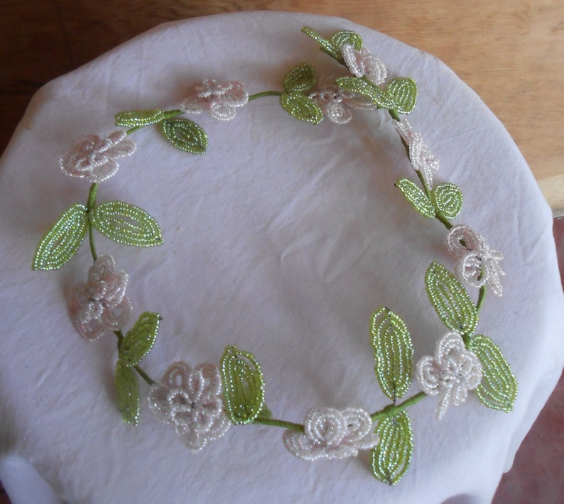 May include: A white and green beaded garland with white flowers and green leaves. The garland is made of small beads and is strung together in a circular shape.
