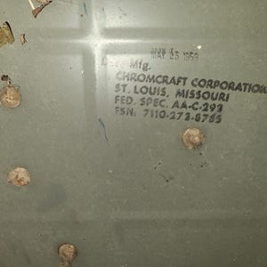 May include: Close-up of a faded, olive-green metal surface with visible wear and tear. The text "MAY 25 1959" is printed at the top, with additional text below indicating "CHROMCRAFT CORPORATION, ST. LOUIS, MISSOURI". Several rivets are visible.