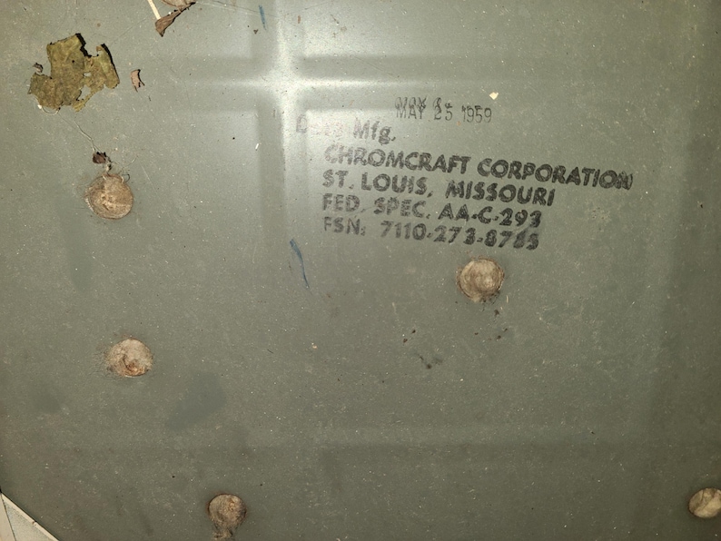 May include: Close-up of a faded, olive-green metal surface with visible wear and tear. The text "MAY 25 1959" is printed at the top, with additional text below indicating "CHROMCRAFT CORPORATION, ST. LOUIS, MISSOURI". Several rivets are visible.