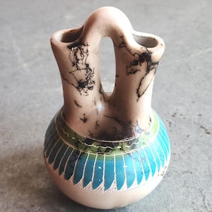 May include: A beige ceramic vase with a double spout and a rounded base. The vase features a turquoise and green band with a geometric pattern. The upper portion has black markings, adding a unique touch to this decorative piece.