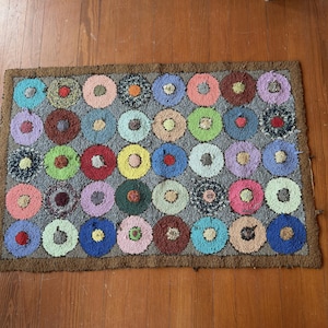 May include: A rectangular rug featuring a grid of multi-colored circular patterns on a grey background. Each circle contains a smaller contrasting center. The rug is bordered by a thick brown frame, showcasing a textured, looped pile construction.