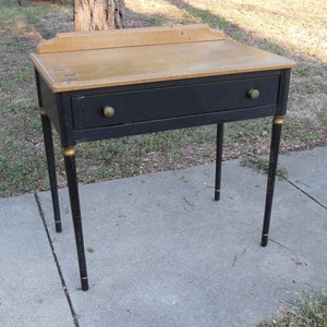 Pu&ograve; includere: Una scrivania in legno nero con un solo cassetto e dettagli dorati sulle gambe. Il piano della scrivania &egrave; in legno chiaro.