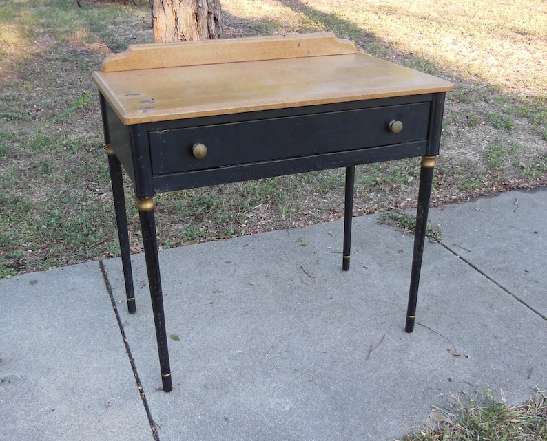 Pu&ograve; includere: Una scrivania in legno nero con un solo cassetto e dettagli dorati sulle gambe. Il piano della scrivania &egrave; in legno chiaro.