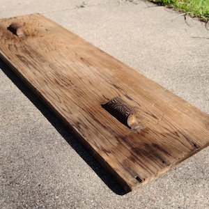 May include: A weathered, rectangular wooden board with two ornate, metal handles. The wood has a rich, brown color with visible grain and dark streaks. The handles are a dark, aged metal, adding to the rustic aesthetic.