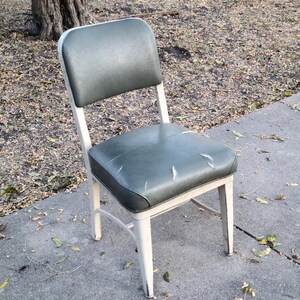 May include: A vintage chair with a white metal frame and a worn, dark green seat and backrest. The seat shows signs of wear. The chair is positioned outdoors on a concrete surface.