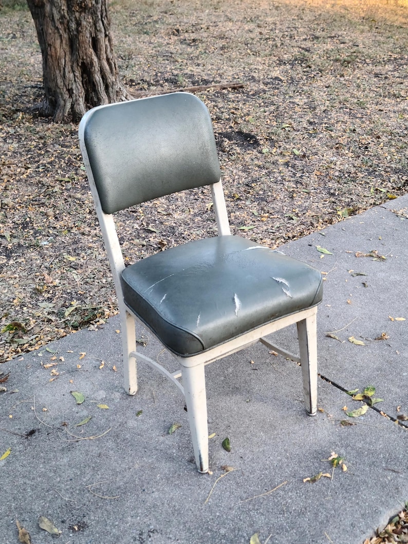 May include: A vintage chair with a white metal frame and a worn, dark green seat and backrest. The seat shows signs of wear. The chair is positioned outdoors on a concrete surface.