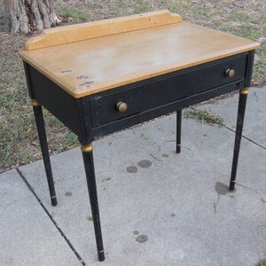 Pu&ograve; includere: Una scrivania nera con un piano in legno chiaro e dettagli dorati. La scrivania ha un cassetto e quattro gambe.