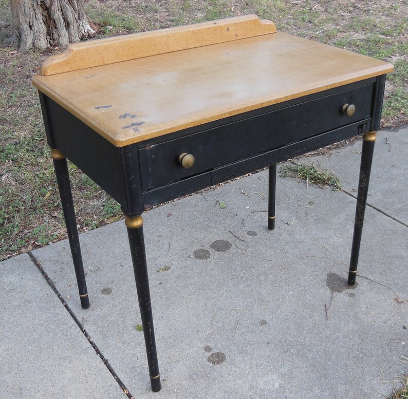 Pu&ograve; includere: Una scrivania nera con un piano in legno chiaro e dettagli dorati. La scrivania ha un cassetto e quattro gambe.