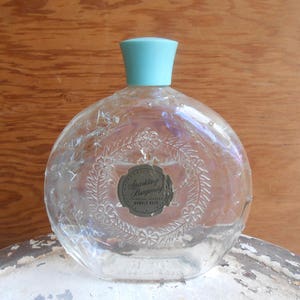 May include: A clear glass bottle with a light blue plastic lid and a label that reads "Sparkling Burgundy Bubble Bath". The bottle has a textured surface with a floral design.
