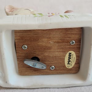 May include: A white ceramic music box with a wooden base. The music box has a decorative design on top and a wind-up key on the bottom. The label reads "MANN MCMLXXXII Japan".