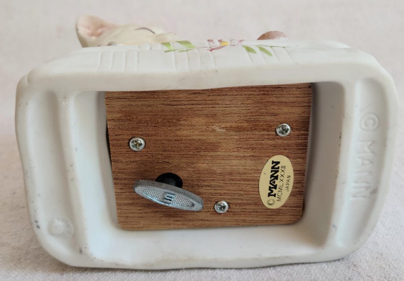 May include: A white ceramic music box with a wooden base. The music box has a decorative design on top and a wind-up key on the bottom. The label reads "MANN MCMLXXXII Japan".