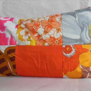 May include: Rectangular patchwork pillow featuring a variety of floral patterns in shades of pink, orange, yellow, and gray. The pillow has a solid orange center panel and is likely used for home decor.