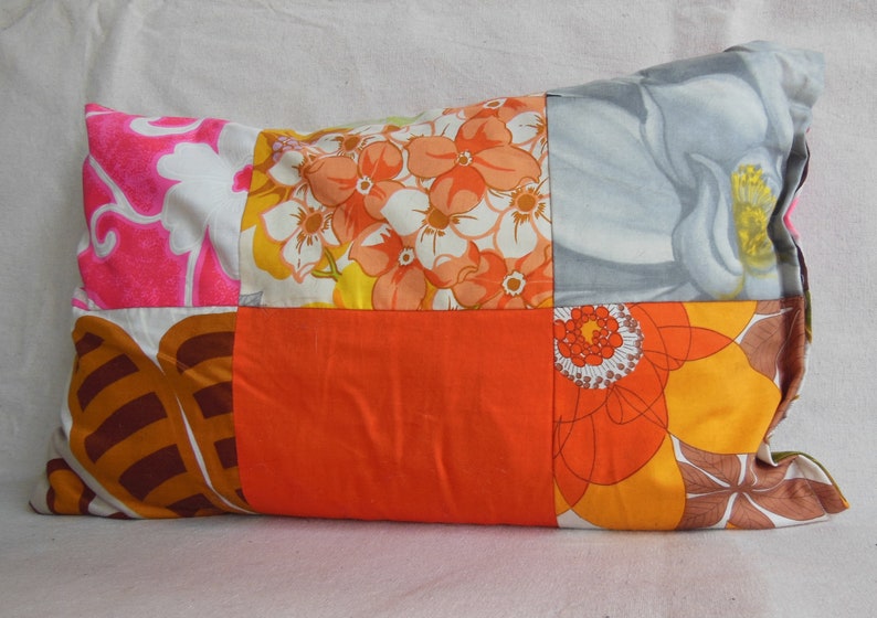 May include: Rectangular patchwork pillow featuring a variety of floral patterns in shades of pink, orange, yellow, and gray. The pillow has a solid orange center panel and is likely used for home decor.