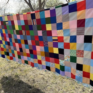 May include: A colorful patchwork quilt, composed of numerous fabric squares in a variety of colors and patterns. The quilt is displayed outdoors, hanging from a line, with a grassy background. The squares include solid colors and patterned fabrics.