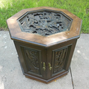 May include: Octagonal wooden table with a carved top and two doors. The top is made of dark wood and has a carved design of a scene with trees and animals. The doors are also made of dark wood and have carved designs.