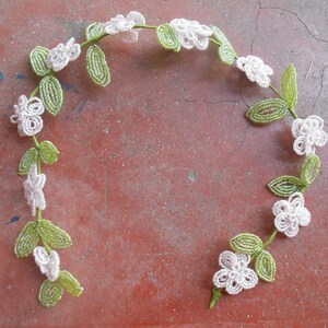 May include: A beaded garland with white flowers and green leaves. The garland is made of small beads and is strung together to form a circle.