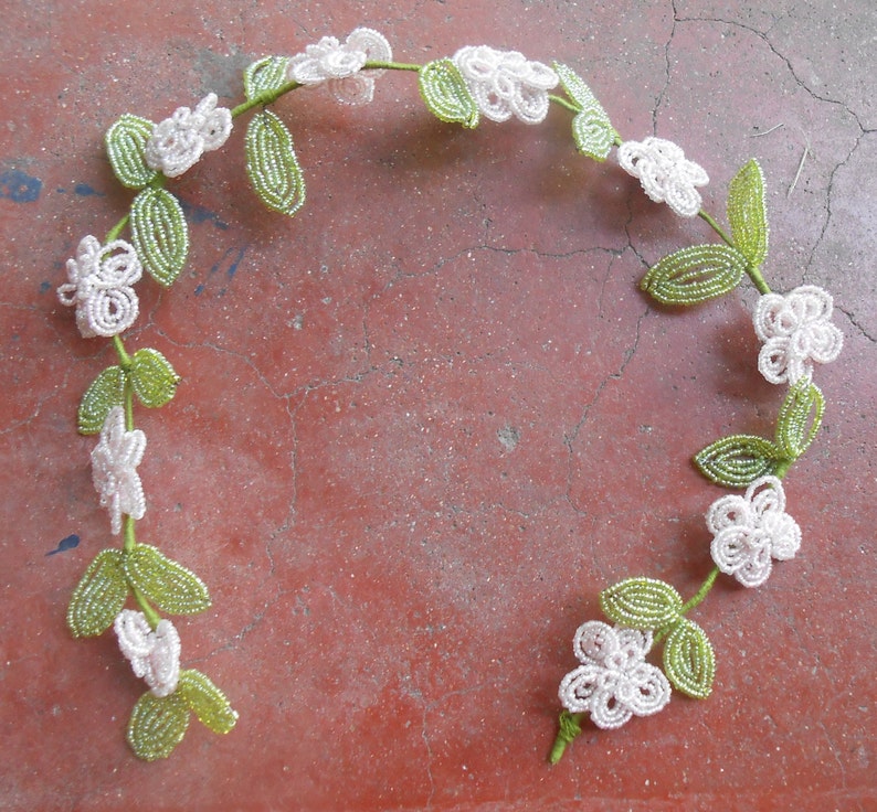 May include: A beaded garland with white flowers and green leaves. The garland is made of small beads and is strung together to form a circle.