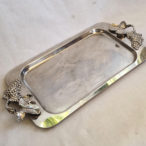 May include: A silver serving tray with grapevine-shaped handles. The tray is rectangular and has a smooth, polished surface.