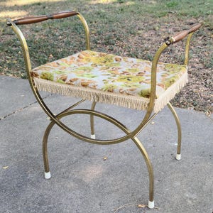 May include: Vintage vanity stool with a gold-tone metal frame and a floral patterned seat. The seat is upholstered with a cream-colored fabric featuring yellow and green floral designs, and trimmed with a beige fringe. The stool has wooden armrests.