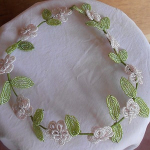 May include: A delicate beaded wreath with white flowers and green leaves. The wreath is made of small beads and is perfect for adding a touch of elegance to any home decor.