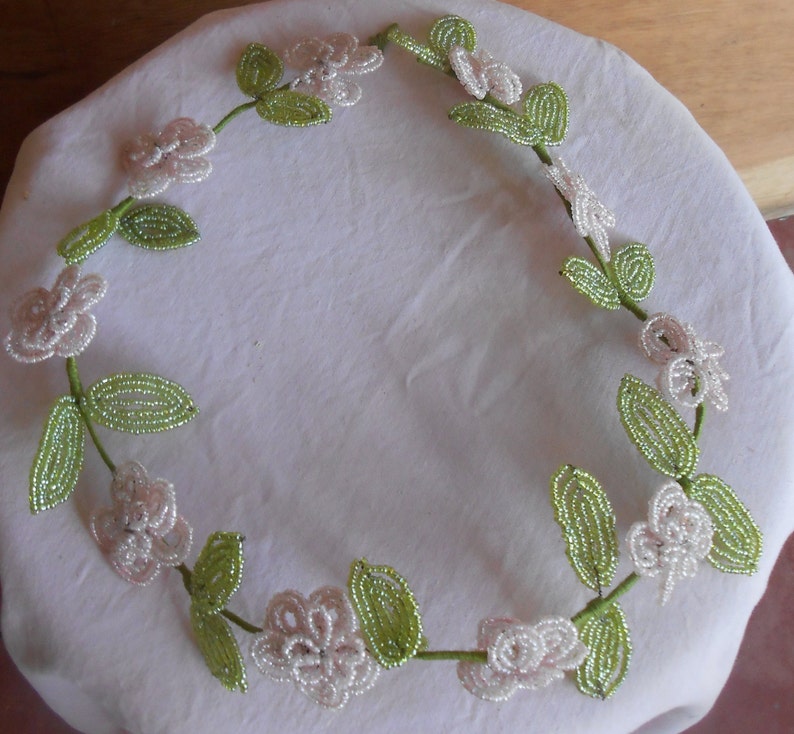 May include: A delicate beaded wreath with white flowers and green leaves. The wreath is made of small beads and is perfect for adding a touch of elegance to any home decor.