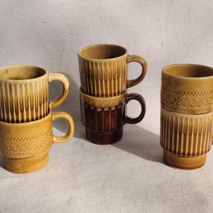 May include: Set of six brown and beige ceramic mugs with a geometric pattern. The mugs are stacked in pairs and have a handle on each side.