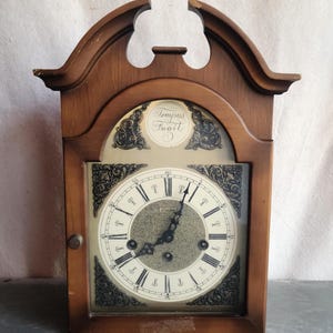 May include: A wooden wall clock with a light-colored face and Roman numerals. The clock has a dark brown wooden frame and decorative scrollwork around the face. The text "Tempus Fugit" is visible on the clock face.