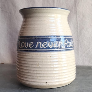May include: Cream-colored ceramic vase with a blue band featuring the words "Love never fails." The vase has a ribbed texture and a slightly flared top. A decorative piece for the home.