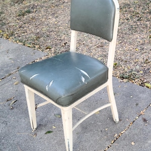 May include: A vintage chair with a distressed, cream-coloured metal frame. The seat and back are upholstered in worn, olive-green vinyl. The chair has a simple, functional design, suitable for vintage or industrial decor.