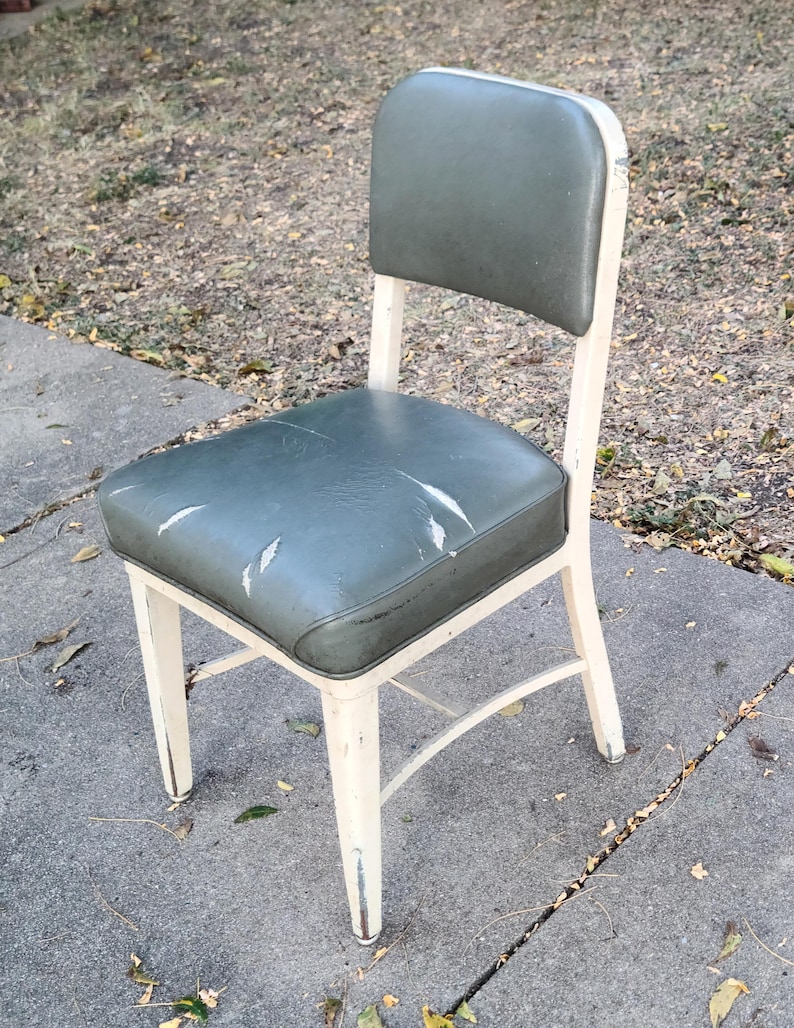 May include: A vintage chair with a distressed, cream-coloured metal frame. The seat and back are upholstered in worn, olive-green vinyl. The chair has a simple, functional design, suitable for vintage or industrial decor.