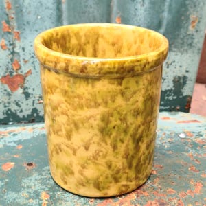 May include: A cylindrical ceramic container with a speckled yellow and green glaze. The container has a slightly flared rim and a smooth, glossy finish. The overall design is rustic and vintage.