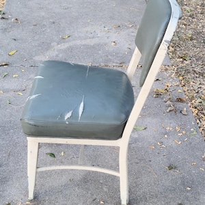 May include: A vintage metal chair featuring a worn olive-green vinyl seat and back. The frame is painted off-white, with signs of age. The seat shows significant wear and tear, indicating it's been used. The chair is likely an antique.