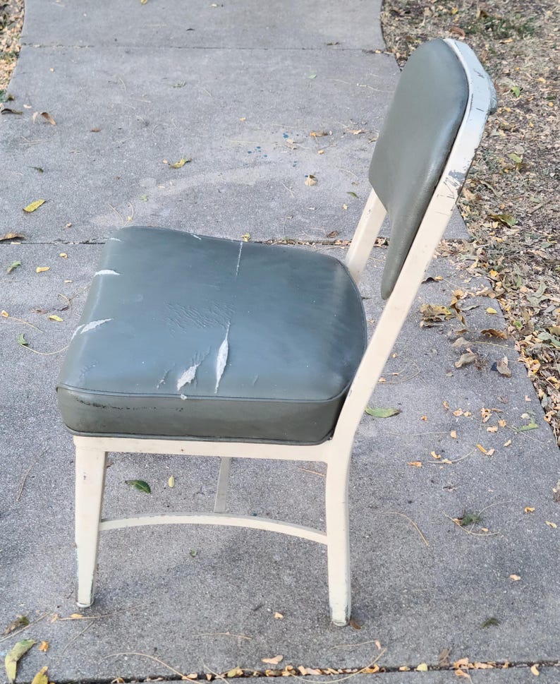 May include: A vintage metal chair featuring a worn olive-green vinyl seat and back. The frame is painted off-white, with signs of age. The seat shows significant wear and tear, indicating it's been used. The chair is likely an antique.