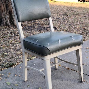 May include: An old chair with a worn, dark green seat and back. The seat shows signs of damage. The frame is painted off-white, with signs of age. The chair is outdoors on a concrete surface.
