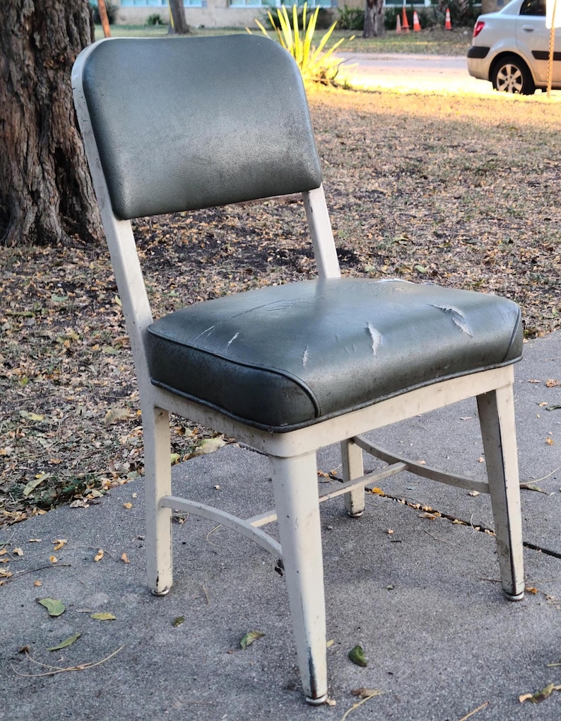 May include: An old chair with a worn, dark green seat and back. The seat shows signs of damage. The frame is painted off-white, with signs of age. The chair is outdoors on a concrete surface.