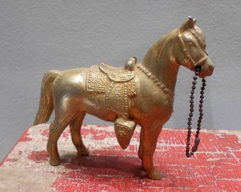 May include: A gold-colored metal horse figurine with a saddle and a chain. The horse is standing on a red wooden surface.