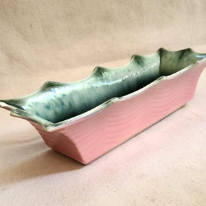 May include: A rectangular, pink ceramic planter with a wavy texture. The rim and interior are glazed in a mottled green-blue color. The planter has a unique, scalloped edge design, adding a decorative touch to the piece.