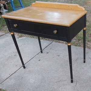Pu&ograve; includere: Un tavolo in legno nero con un solo cassetto e dettagli dorati sulle gambe. Il piano del tavolo &egrave; in legno marrone chiaro.