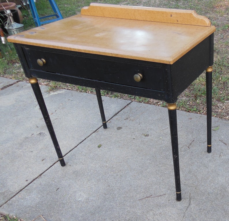 Pu&ograve; includere: Un tavolo in legno nero con un solo cassetto e dettagli dorati sulle gambe. Il piano del tavolo &egrave; in legno marrone chiaro.