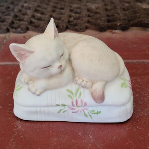 May include: A cream-colored ceramic cat figurine sleeping on a white pillow. The pillow is decorated with pink and green floral designs. The cat is curled up, with its eyes closed and its tail resting on the pillow.